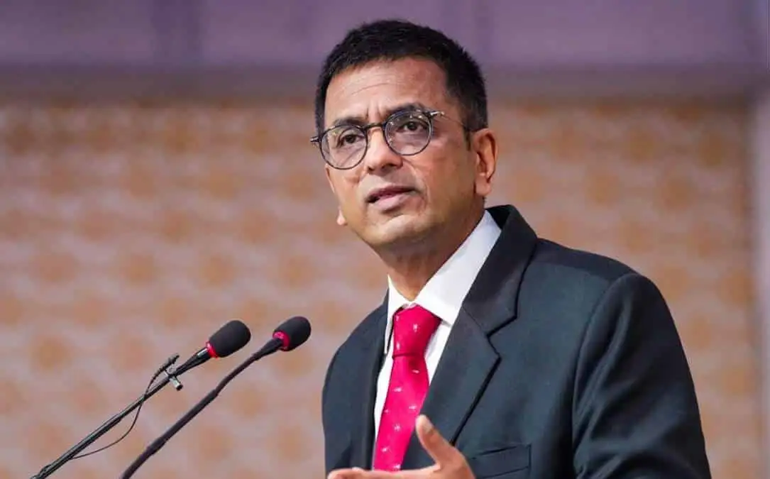 A man in a suit and red tie speaks at a podium with two microphones. The background is out of focus with a warm, patterned design.