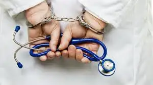 Hands in handcuffs holding a blue stethoscope. Person wears a white coat, suggesting a medical setting. Tense mood. No text visible.