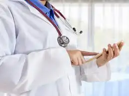 Doctor in a white coat and stethoscope uses a tablet. Bright, airy room with large windows in the background, conveying a professional atmosphere.
