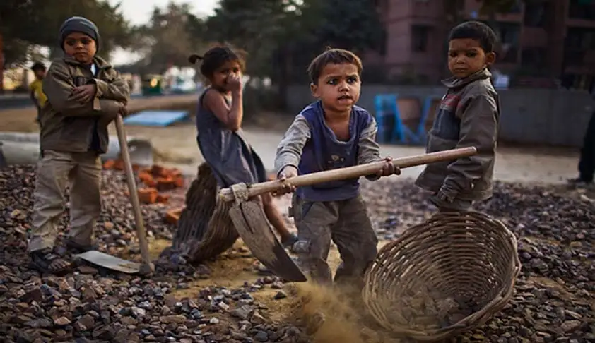 Children working outdoors with shovels, baskets, and dirt. They appear focused and serious. Trees and buildings in the blurred background.