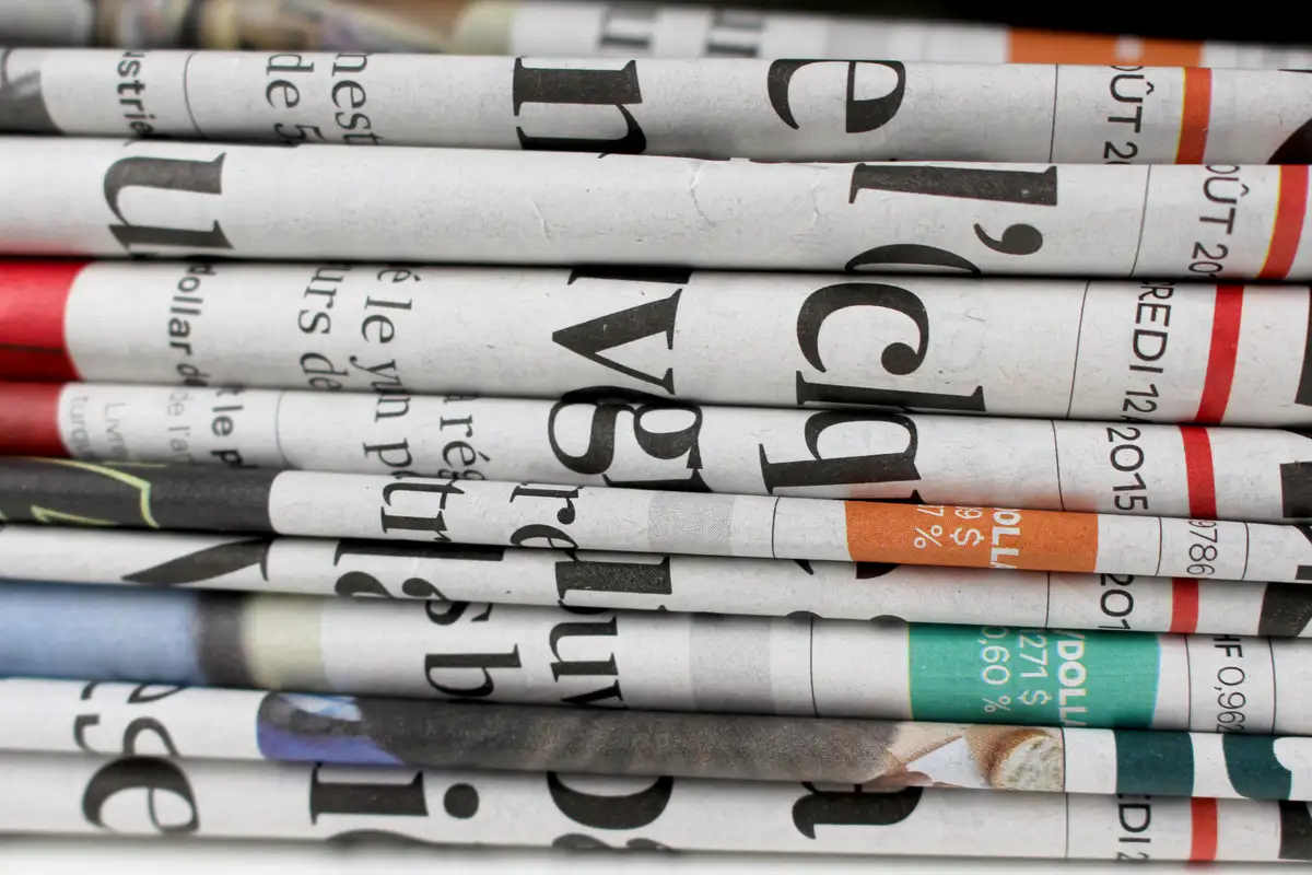 Stack of folded newspapers with bold headlines. Red, orange, and teal sections visible. Close-up, focused on text and colors.