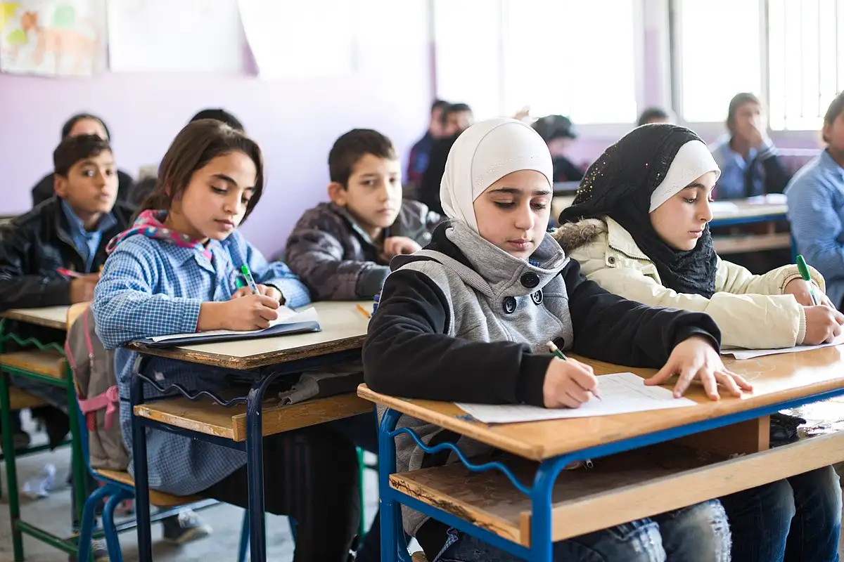 Students seated at desks in a classroom, writing attentively. Bright light from a window. Focused atmosphere with pastel walls.
