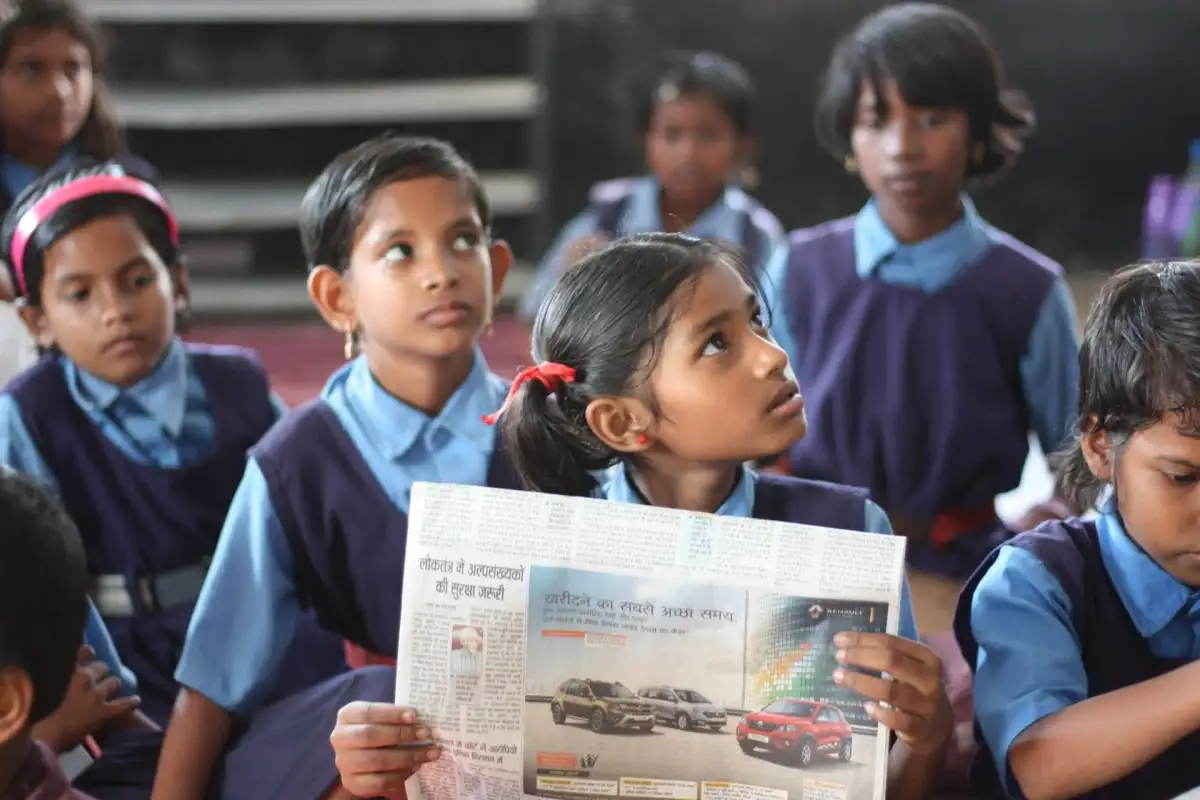 Young students in uniforms sit attentively in class. A girl holds a newspaper; others look on curiously. Background shows blurred peers.